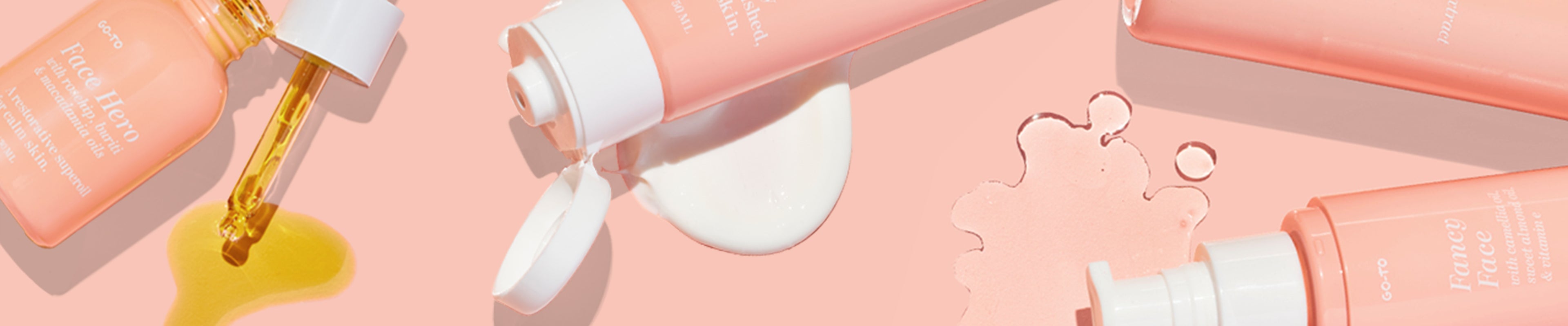 Several pink skincare bottles and a dropper with golden oil lie on a peach background. One bottle spills a white liquid, creating small puddles next to the products.