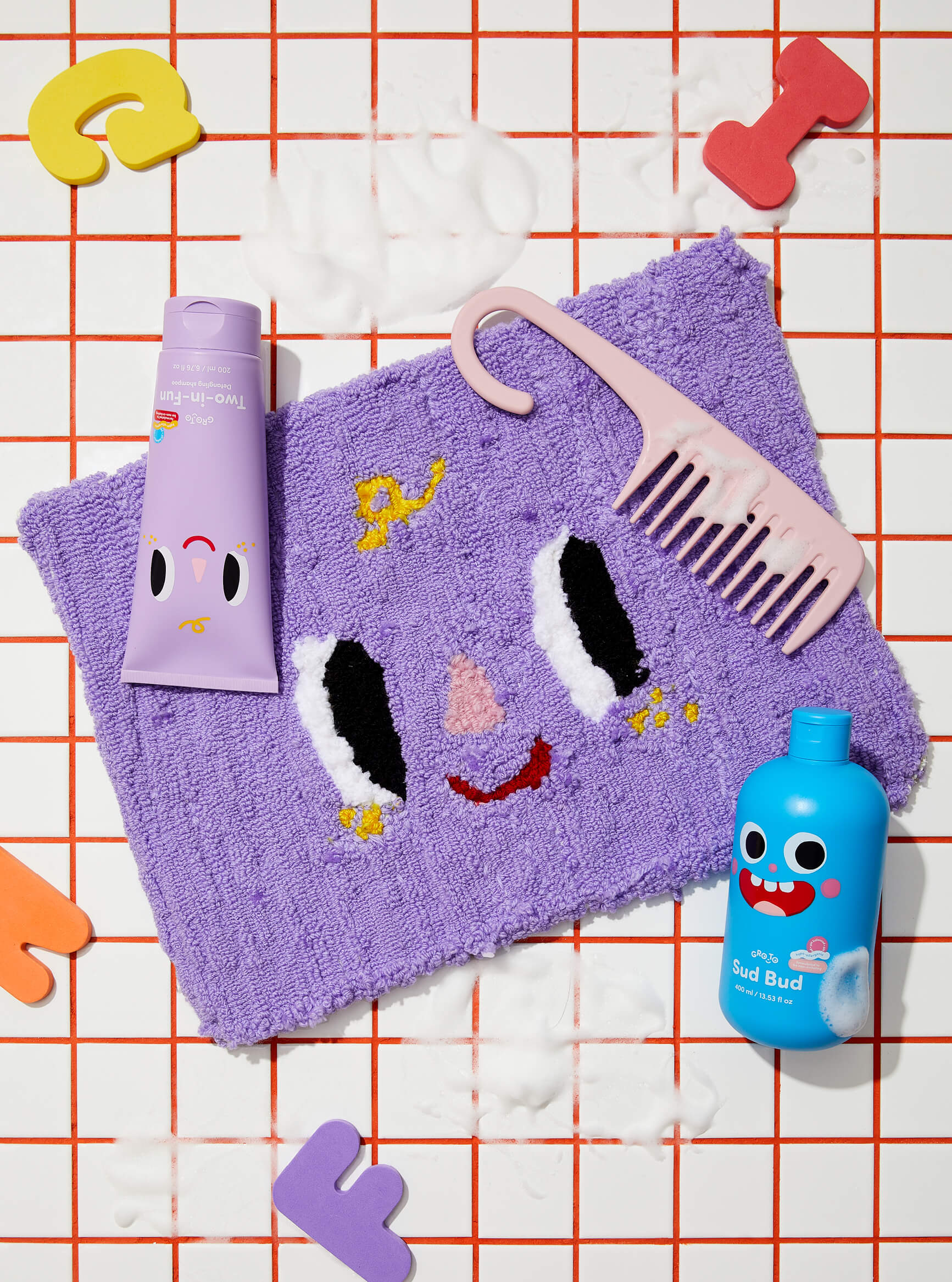 A purple towel with a cute face design, a pink wide-tooth comb, purple and blue bottles with smiling faces, and colorful foam letters on a red grid tiled surface with soap suds.