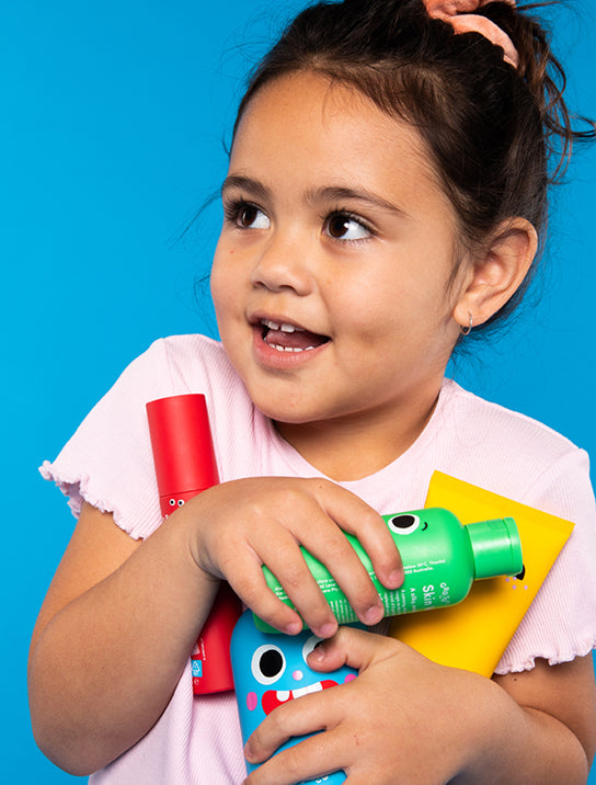 A young girl in a light pink shirt smiles while holding colorful bottles and containers with playful faces against a bright blue background.