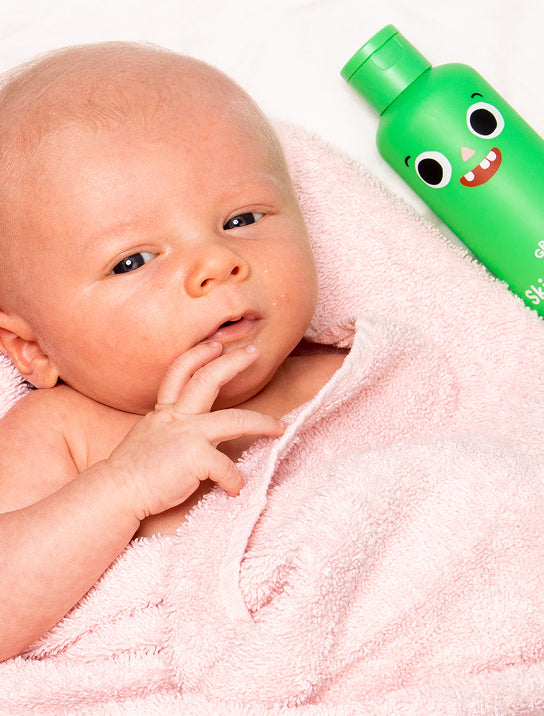 A baby wrapped in a soft pink towel lies next to a green bottle with a smiling cartoon face on it. The baby looks up with one hand near its mouth.