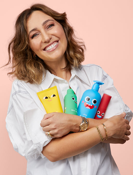 A smiling woman in a white shirt hugs four colorful skincare products with cartoon faces on their packaging against a pink background.