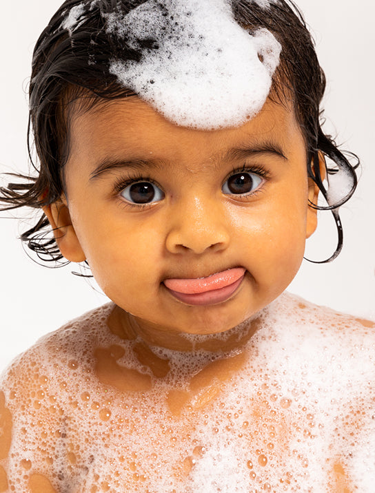 A young child with wet hair and big brown eyes has a playful expression, sticking out their tongue. The child is covered in soapy bubbles, including a dollop of foam on their head, against a plain white background.