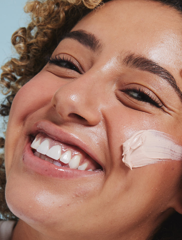 A person with curly hair smiles warmly, showing their teeth. They have a swipe of cream on their cheek, and their skin looks smooth and radiant. The background is a light blue color.