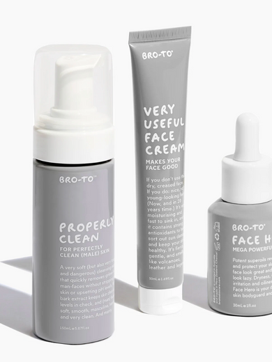 Three skincare products from the brand Bro-To are arranged on a white surface: a foaming cleanser, a tube of face cream, and a dropper bottle, all in minimalist gray packaging with white text.