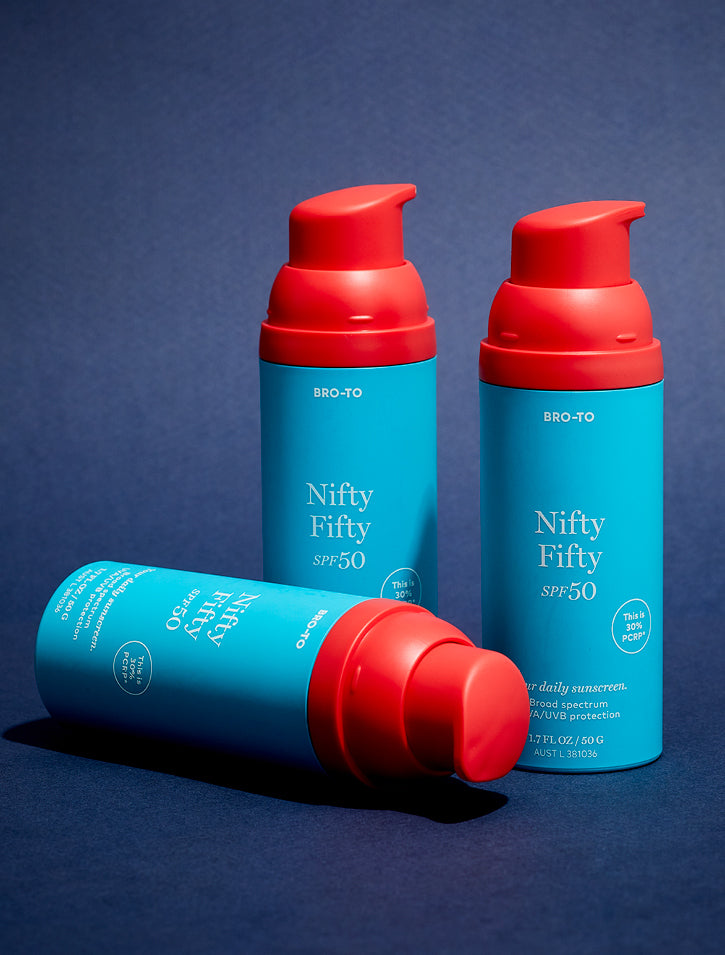 Three blue bottles of BRO-TO Nifty Fifty SPF 50 sunscreen with red pumps, two standing upright and one lying on its side, set against a dark blue background.