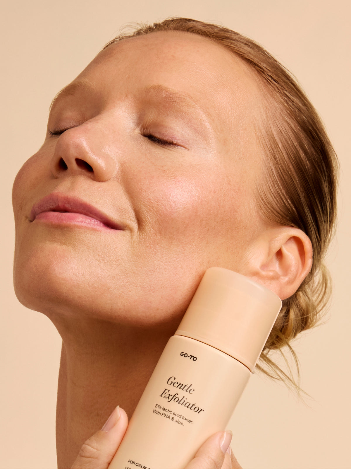 Go-To Skincare Gentle Exfoliator bottle held against a womans neck as she demonstrates a soothing facial exfoliation routine in warm natural light