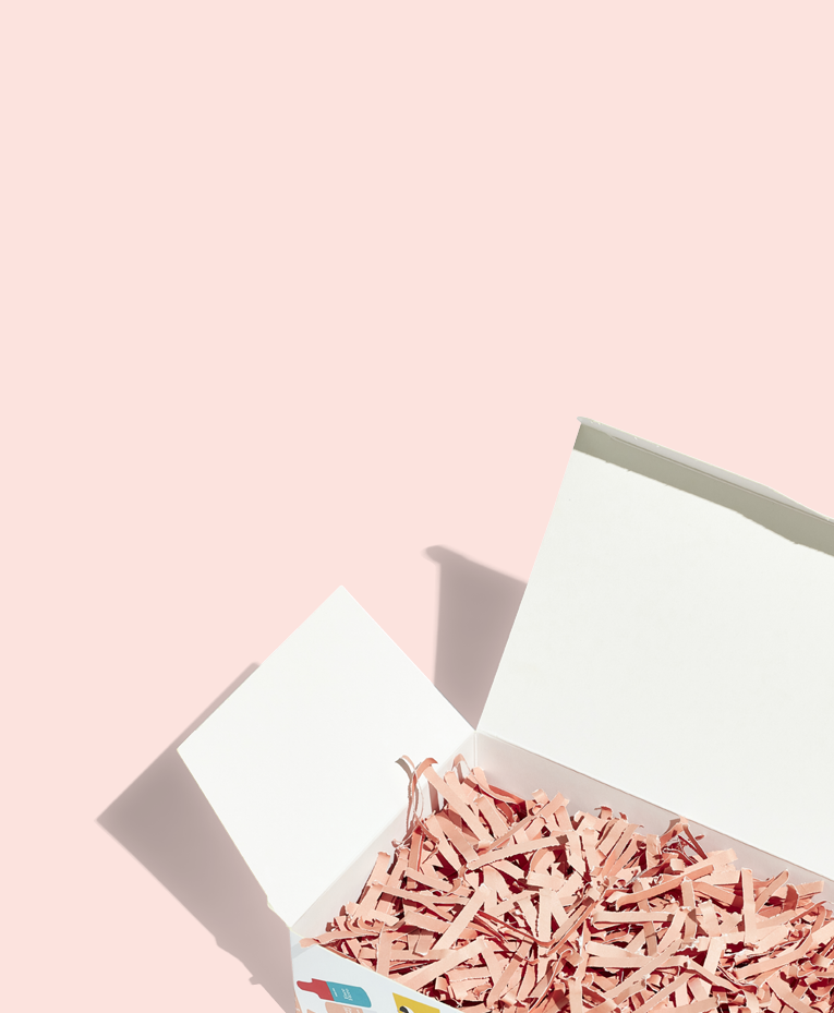 A white open box filled with pink shredded paper sits on a pink surface, casting a shadow. The image has a minimal, pastel aesthetic.