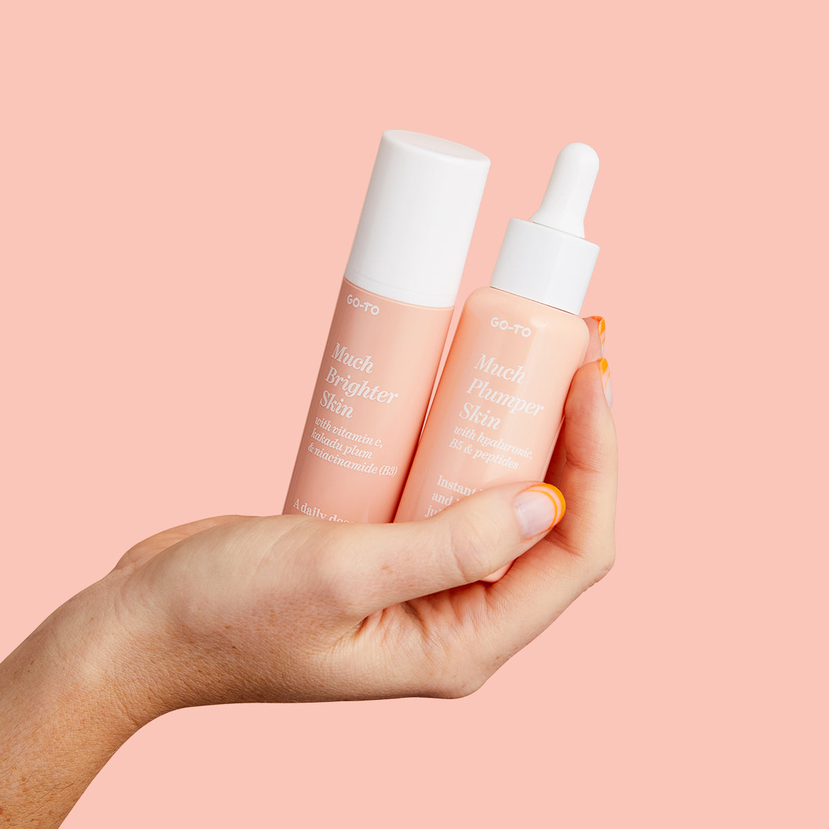 A hand holds two peach-colored skincare products with white labels and caps against a pastel pink background. One bottle has a dropper, and both are branded GO-TO.