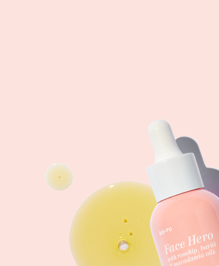 A bottle of Go-To Face Hero oil with a white dropper cap is shown next to drops and a large smear of yellow face oil on a light pink background.