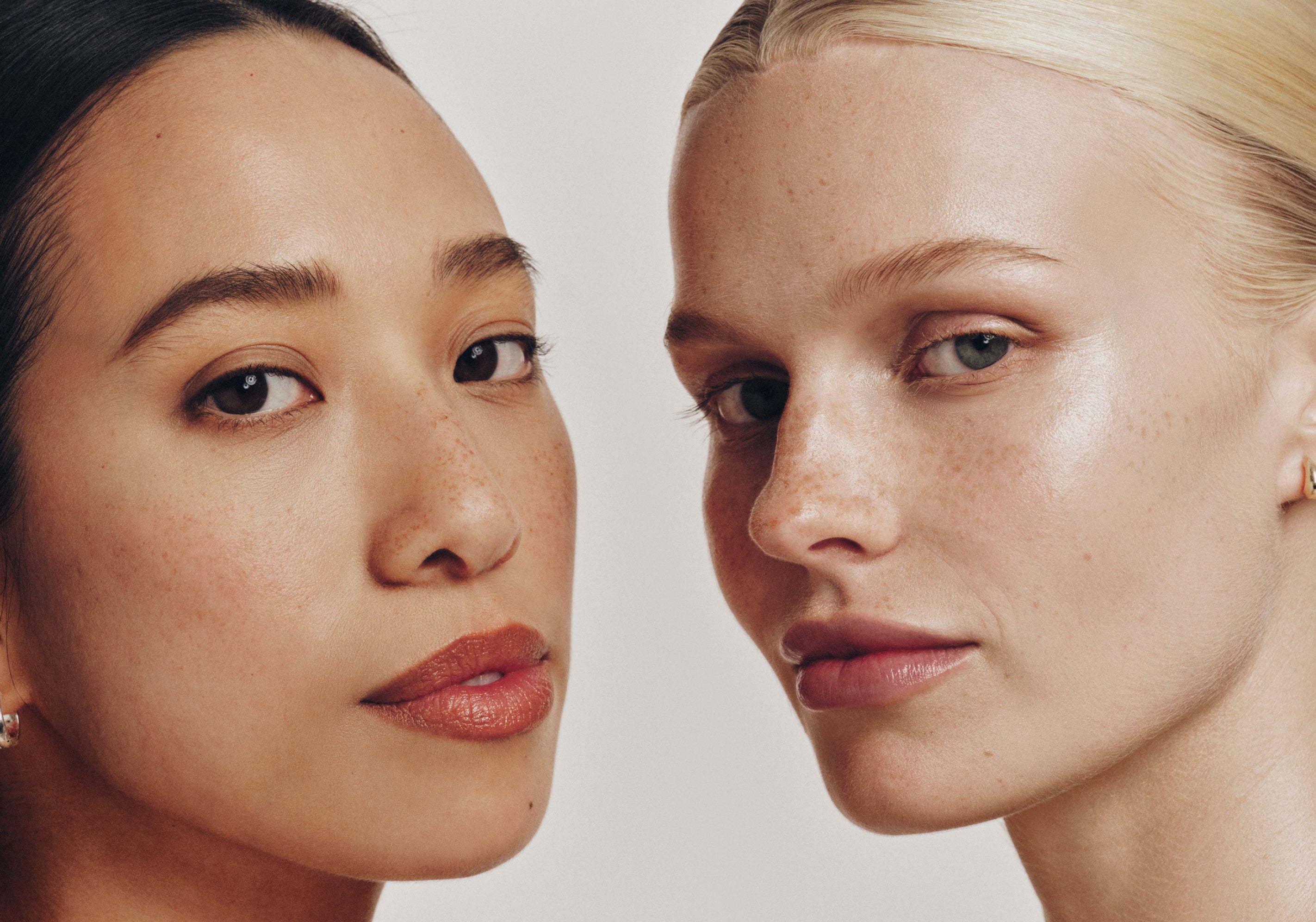 Close-up of two women with clear, glowing skin and minimal makeup, standing side by side against a neutral background. One has dark hair and warm undertones; the other has blonde hair and cool undertones.