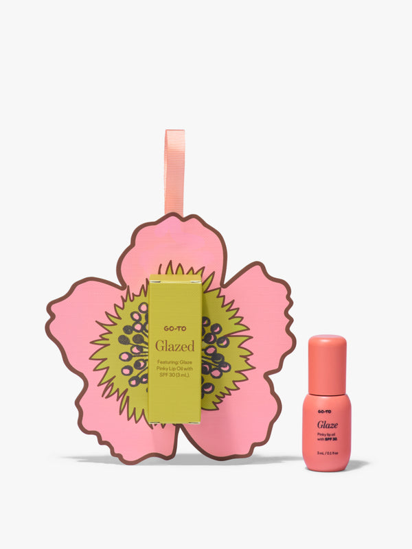 Go To Skincare Glazed tinted SPF lip oil in a pink tube beside its yellow box against an illustrated pink flower backdrop