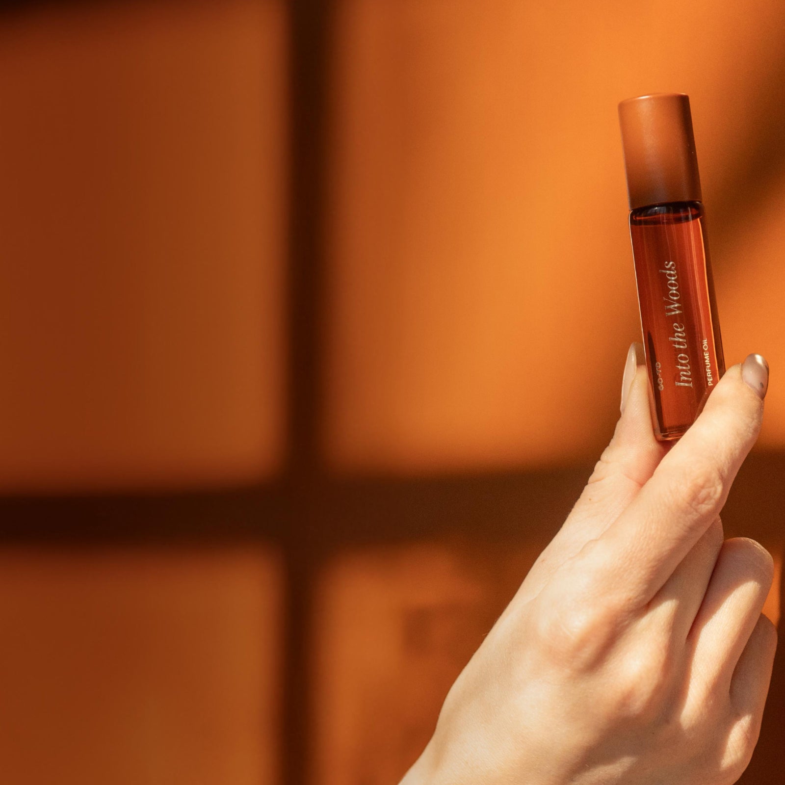 A hand holds up a small amber bottle labeled Into the Woods against a warm orange background with soft, shadowy window lines.