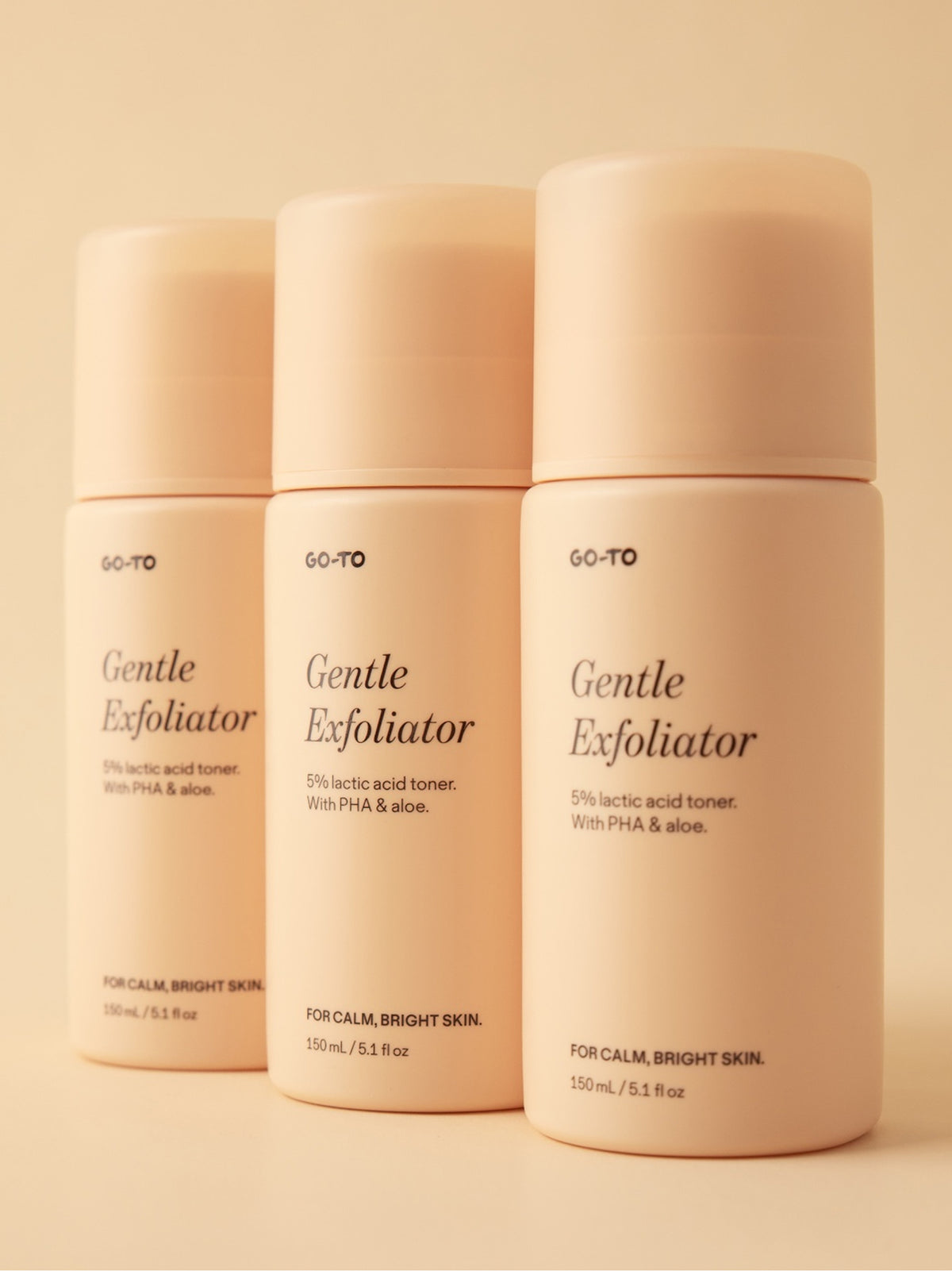 Three Go-To Skincare Gentle Exfoliator bottles with cream caps arranged against a warm beige background. Labels show five percent lactic acid toner with PHA and aloe.